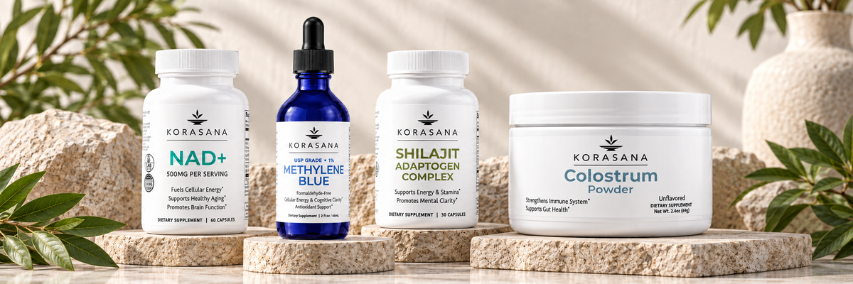 Four Korasana supplement products displayed on a light stone surface with decorative greenery and ceramic vases in the background: NAD+ capsules, Methyln Blue dropper bottle, Shilajit Adaptogen Complex powder, and Colostrum Powder jar.