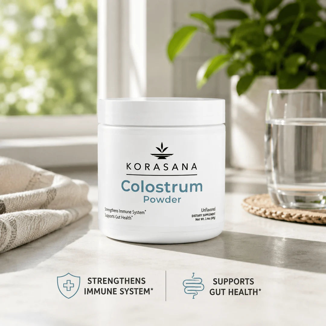 A white jar of KORASANA Colostrum Powder sits on a light countertop near a window, with a glass of water, a folded towel, and a potted plant in the background. Text at the bottom reads 'STRENGTHENS IMMUNE SYSTEM' and 'SUPPORTS GUT HEALTH'.