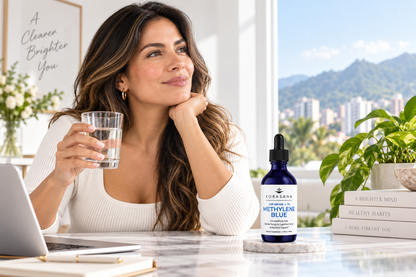 A woman sits at a desk holding a glass of water, with a bottle of Corasana Nutri-Grade Blue dietary supplement in front of her. The setting is a bright, modern room with a window showing a city view and mountains in the background.