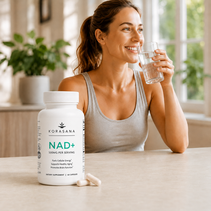 A woman in a gray tank top sits at a table drinking water next to a white bottle of Korasana NAD+ dietary supplements, with two capsules resting in front of the bottle.